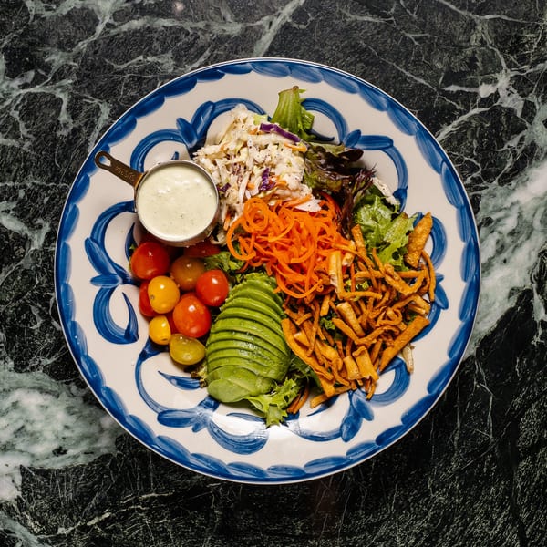 Colorful salad with avocado, cherry tomatoes, shredded carrots, coleslaw, and dressing on a blue and white plate.