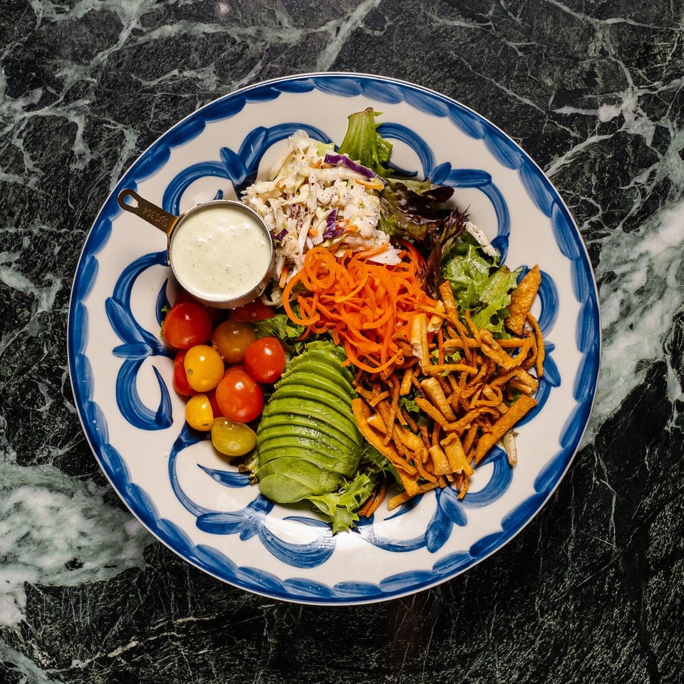 Colorful salad with avocado, cherry tomatoes, shredded carrots, coleslaw, and dressing on a blue and white plate.
