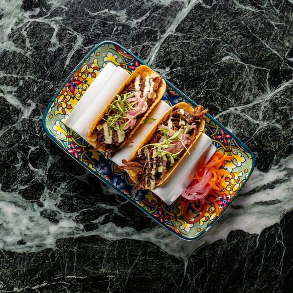 Two tacos with shredded meat, white sauce, and microgreens on a colorful plate with pickled vegetables.