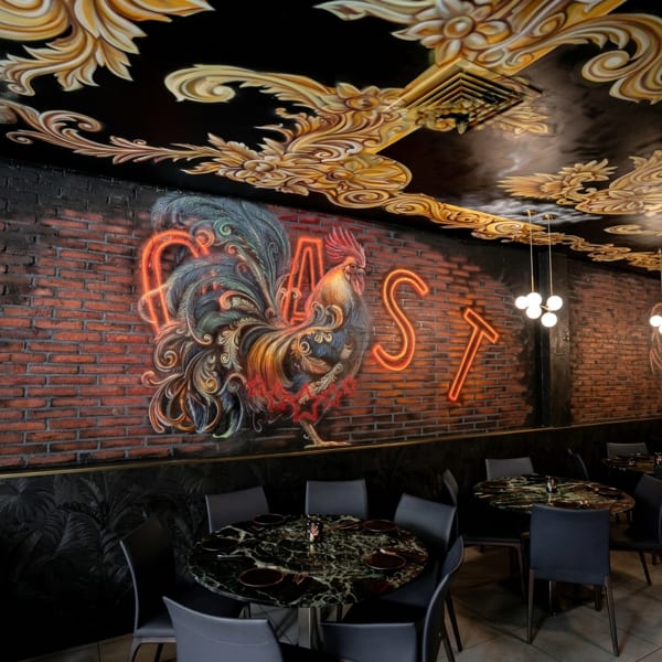 Restaurant interior with a large rooster mural and neon "EAST" sign on a brick wall, with ornate ceiling.