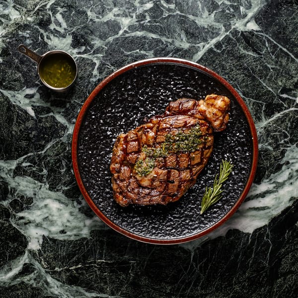Grilled steak with herb butter and rosemary on a black plate, with a side of green sauce.