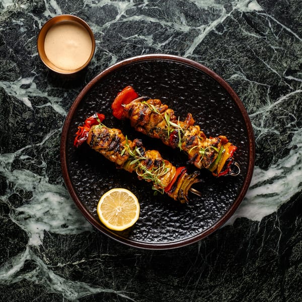 Two grilled chicken and vegetable skewers on a black plate with a lemon slice and dipping sauce.